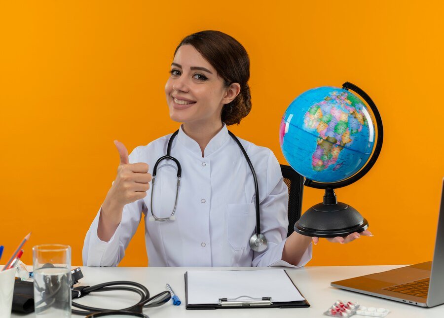 smiling young female doctor wearing medical robe stethoscope sitting desk with medical tools laptop holding globe showing thumb up_141793 58276